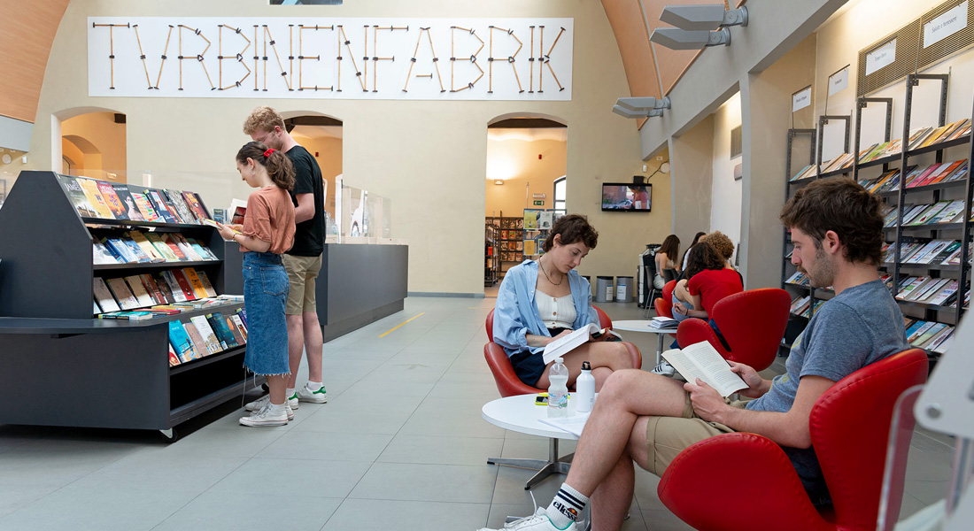 Hall della Biblioteca Lazzerini con alcuni ragazzi seduti a leggere e altri in piedi agli scaffali dei libri